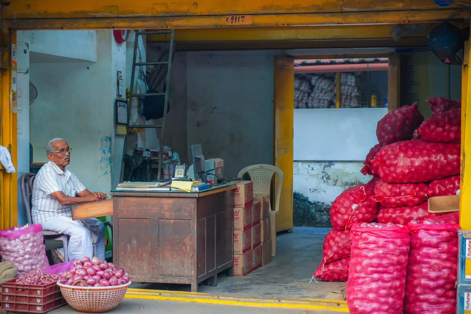 A market shopkeeper surrounded by sacks of red onions and stacked produce boxes